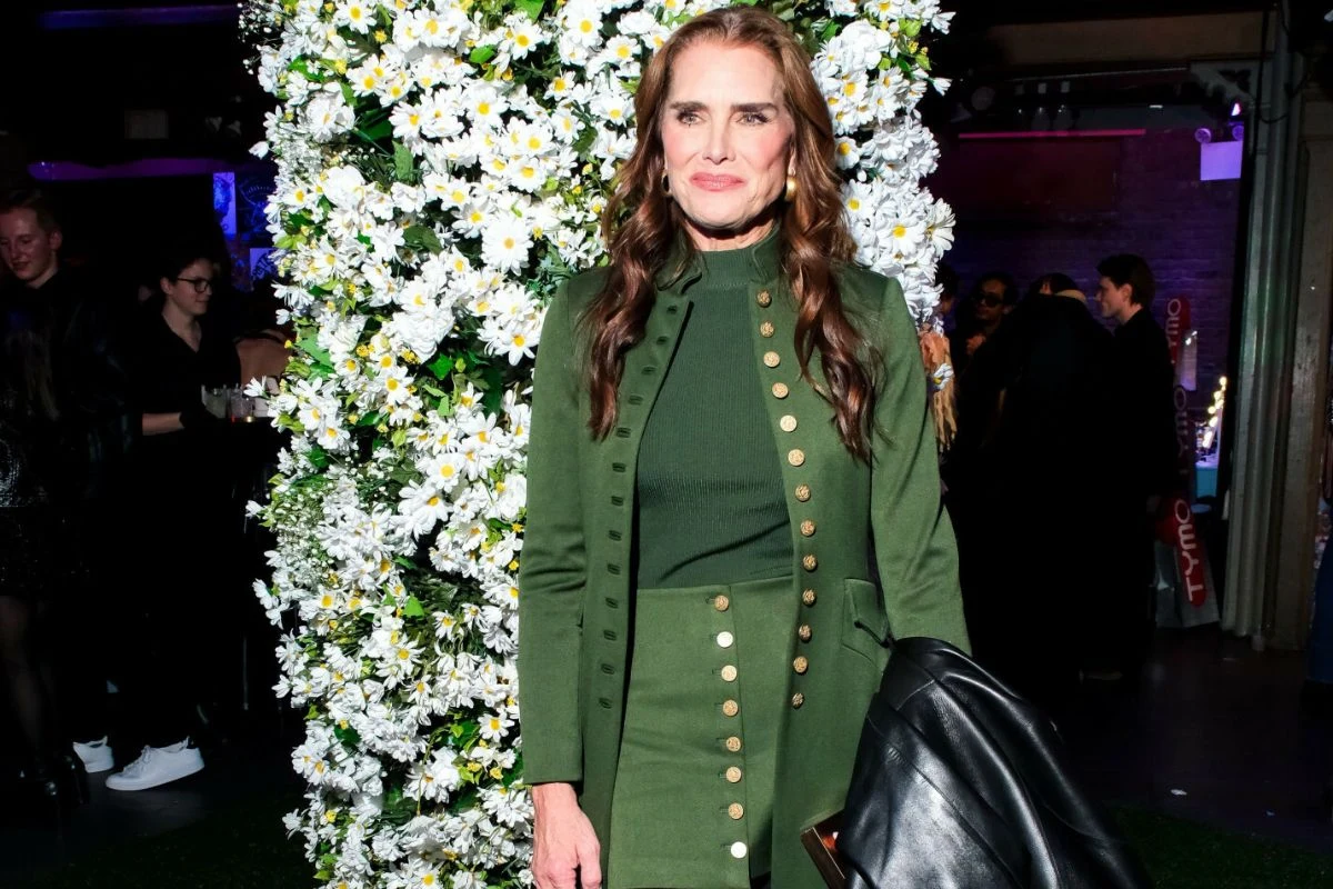 Brooke Shields zachwyca w wielu 60 lat Kobieta w zielonym stroju ze złotymi guzikami stoi na tle ścianki udekorowanej dużą ilością białych kwiatów, w ręku trzyma czarną kurtkę, wokół niej znajdują się inni ludzie w przyciemnionym pomieszczeniu.