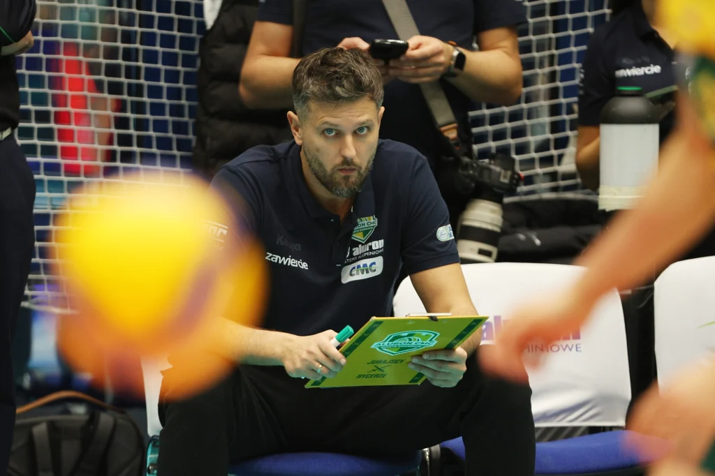 Trener siatkówki siedzący na ławce, trzymający zieloną tabliczkę taktyczną i obserwujący sytuację na boisku. Na pierwszym planie rozmyta piłka oraz części sylwetek zawodników. W tle sprzęt fotograficzny i inne osoby związane z wydarzeniem sportowym.