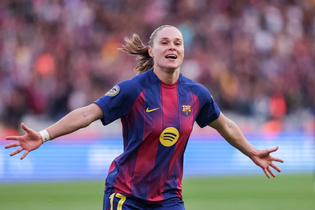 Piłkarka w stroju FC Barcelony z rozłożonymi rękami cieszy się po zdobyciu gola na stadionie, w tle rozmyci kibice.