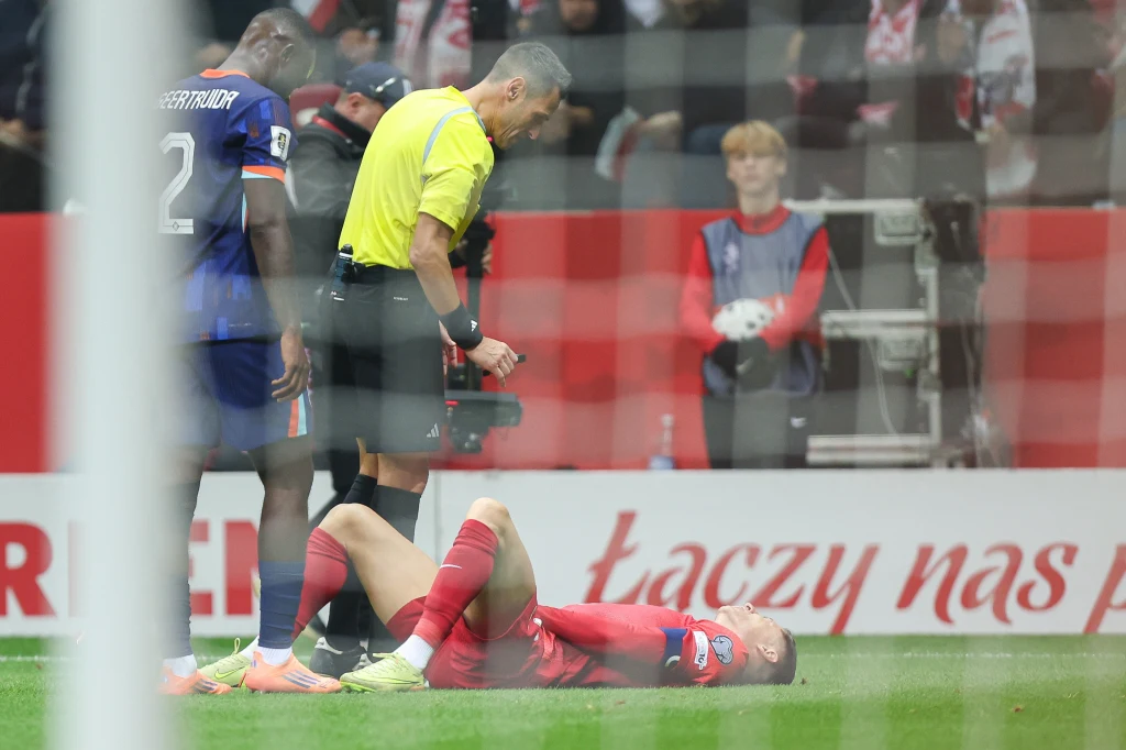 Sebastian Szymański musiał przedwcześnie opuścić boisko w meczu z Holandią Zawodnik w czerwonym stroju leży na murawie podczas meczu piłkarskiego, obok stoi sędzia oraz piłkarz drużyny przeciwnej w granatowym stroju. W tle widoczna publiczność i transparenty.