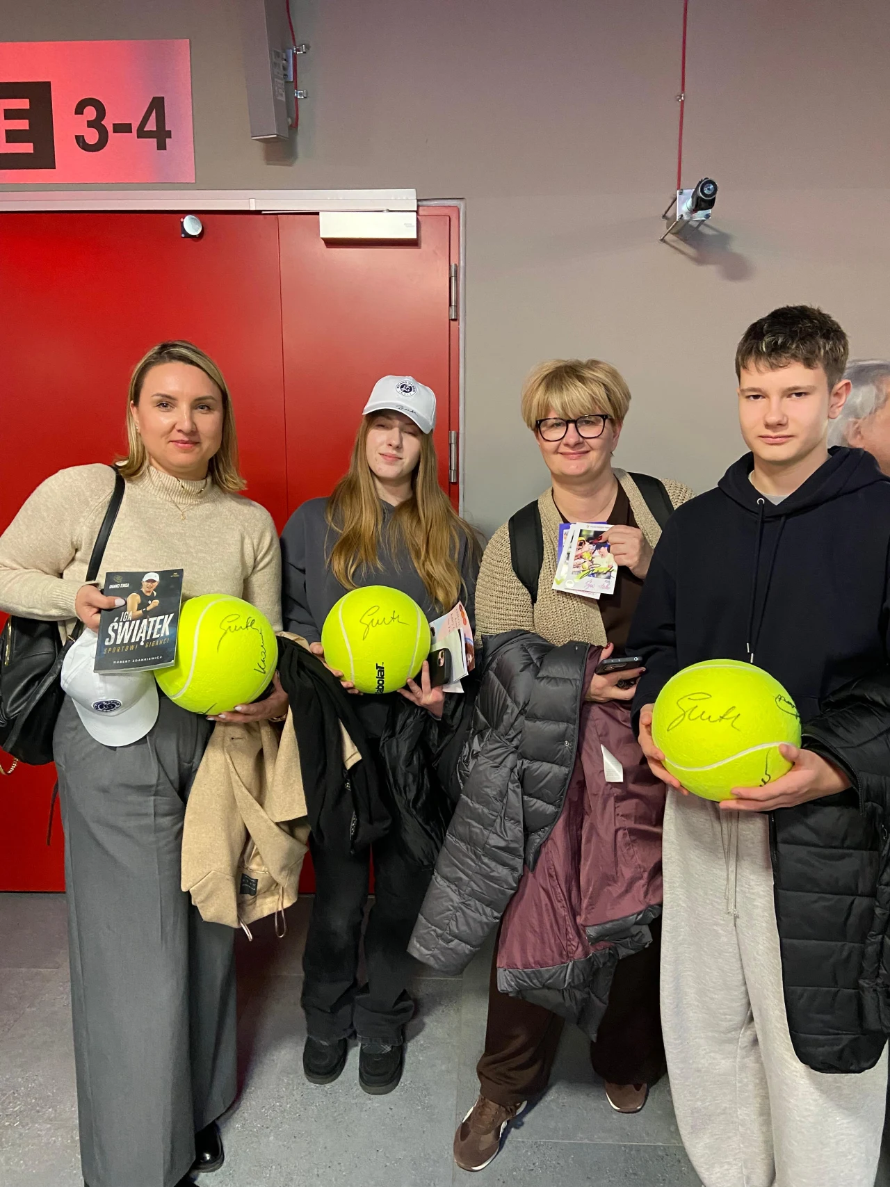 Cztery osoby stojące obok siebie w zamkniętym pomieszczeniu z czerwonymi drzwiami w tle, trzymające duże piłki tenisowe z autografem oraz książki i czapki. Ubrani w zimowe ubrania, wyglądają na zadowolonych, najprawdopodobniej po spotkaniu związanym z ...