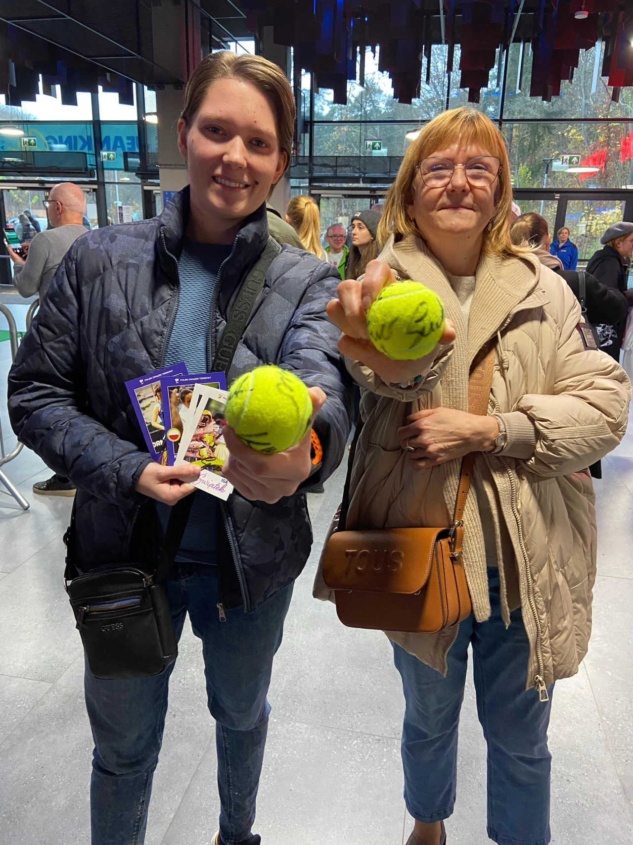 Dwie uśmiechnięte osoby stoją w jasnym wnętrzu, trzymając przed sobą piłki tenisowe z autografami oraz bilety. W tle widoczne są inne osoby oraz szklana ściana z widokiem na zewnątrz.