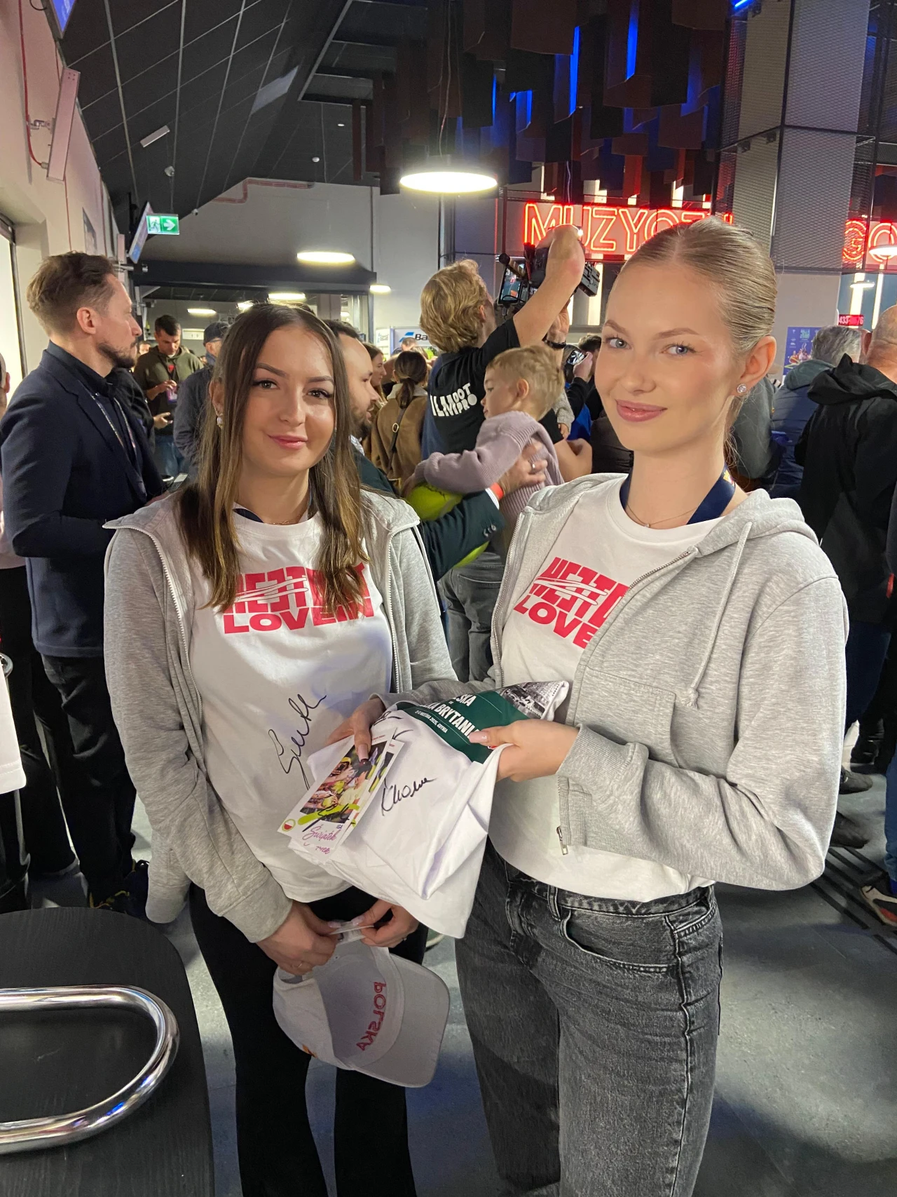 Dwie młode kobiety w białych koszulkach z napisem „Wielka Orkiestra Świątecznej Pomocy” oraz szarych bluzy stoją razem, trzymając podpisane przedmioty i uśmiechając się do aparatu. W tle widać tłum ludzi uczestniczących w wydarzeniu pod świetlnym napis...