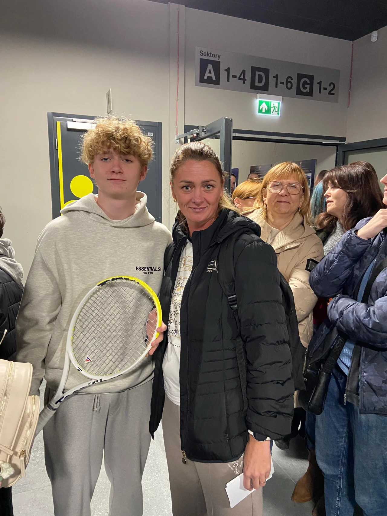 Młody mężczyzna z rakietą tenisową stoi obok kobiety wśród grupy czekających osób w jasnym, zamkniętym pomieszczeniu. W tle widoczne oznaczenia sektorów na ścianie oraz drzwi.