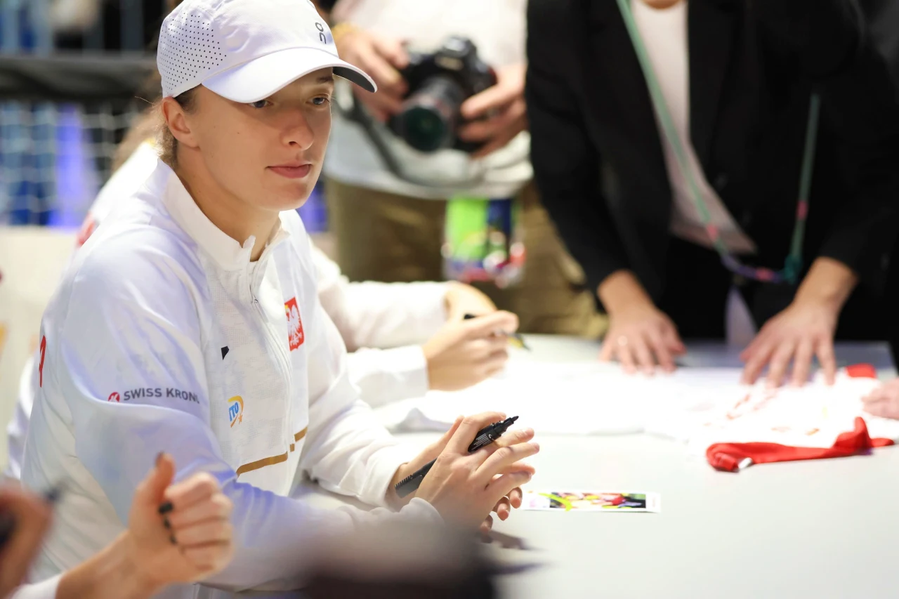 Sportsmenka w białym stroju i czapce z daszkiem siedzi przy stole, w dłoni trzyma marker, prawdopodobnie udzielając autografów. Obok osoby również są skupione na zajęciach, w tle widać fotografa z aparatem.