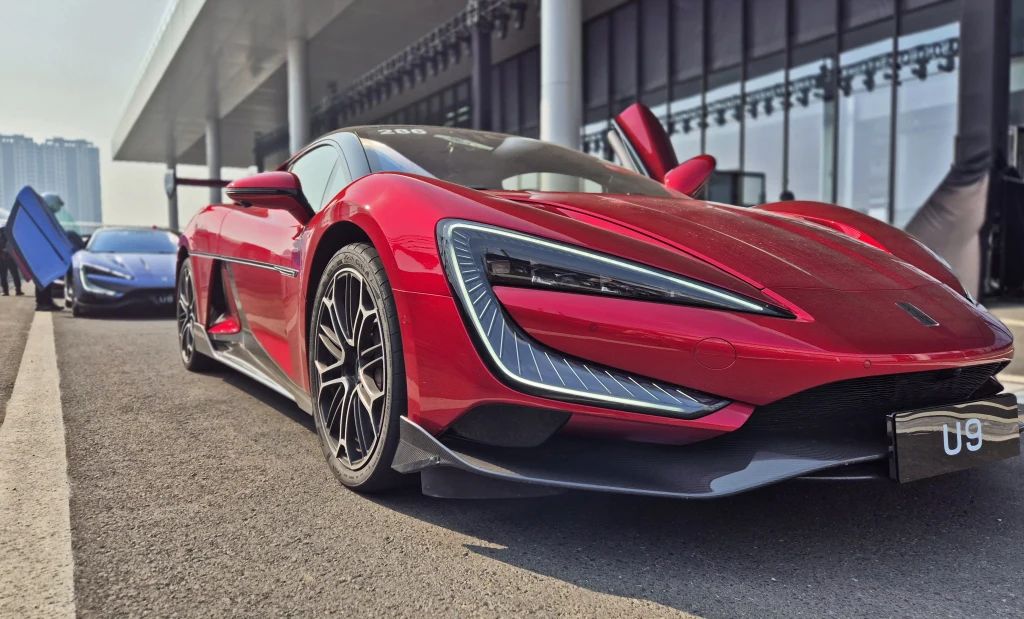 Połacie diodowych pasków nie pozwalają pomylić U9 z innym samochodem Czerwony sportowy samochód z nowoczesnym designem zaparkowany na ulicy, na drugim planie widoczny niebieski pojazd oraz fragmenty nowoczesnego budynku.