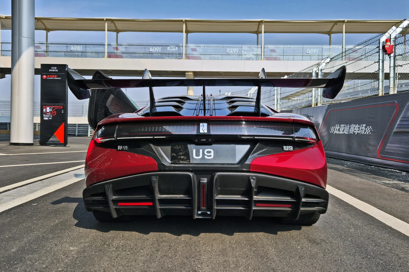 Yangwang U9 nie ma tylnego okna - zamiast niego zastosowano karbonową pokrywę Czerwony sportowy samochód elektryczny z dużym tylnym skrzydłem stoi na torze wyścigowym, widok z tyłu podkreśla aerodynamiczne elementy i masywny dyfuzor, w tle fragment ogrodzenia oraz infrastruktury toru.