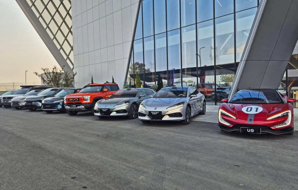 Ośrodek testowy BYD w Zhengzhou Rząd nowoczesnych samochodów ustawionych przed szklanym budynkiem, od aut terenowych po sportowe, z czerwonym, wyścigowym modelem na końcu szeregu.