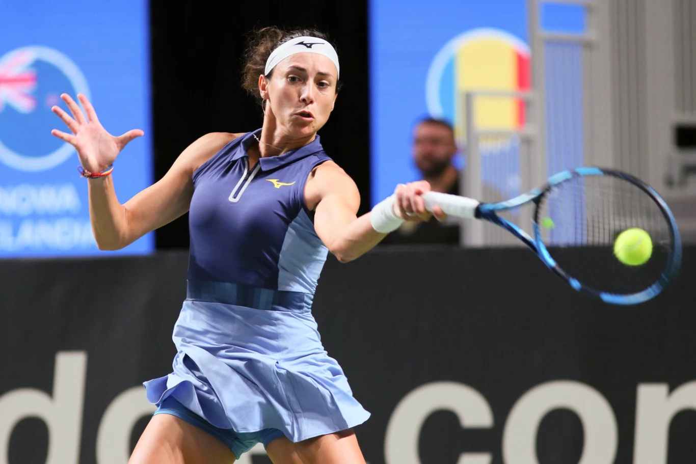 Zawodniczka w sportowym stroju podczas dynamicznego uderzenia piłki tenisowej rakietą na korcie, w tle widoczne rozmazane postacie oraz część tablicy informacyjnej z flagami.