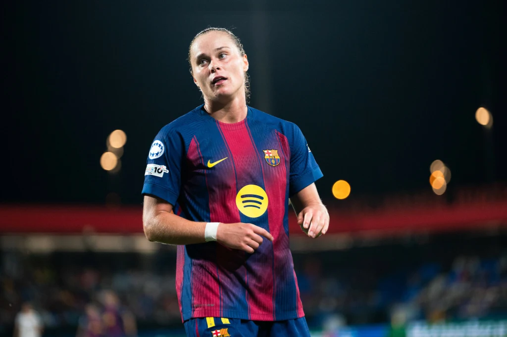 Piłkarka w koszulce FC Barcelony na stadionie podczas meczu, z widocznym logotypem sponsorów na przodzie stroju, tło rozmazane, światła stadionowe w wieczornej scenerii.