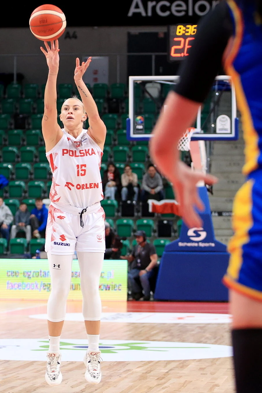 Zawodniczka polskiej reprezentacji koszykówki oddaje rzut w trakcie meczu, ubrana w biały strój z napisem Polska i numerem 15, w tle widoczny fragment boiska, trybuny oraz przeciwniczka w niebiesko-żółtym stroju.