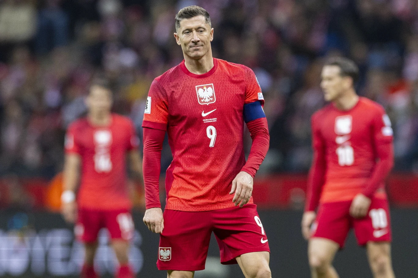 Robert Lewandowski Piłkarz w czerwonym stroju reprezentacji Polski z opaską kapitana na ramieniu stoi na murawie, w tle widoczni są pozostali członkowie drużyny podczas meczu.