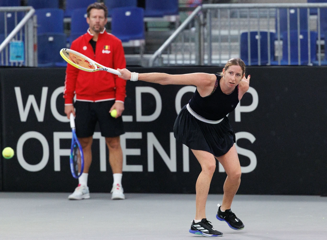 Zawodniczka tenisowa uderza piłkę na korcie, ubrana w czarną sukienkę sportową. W tle trener w czerwonej bluzie obserwuje przebieg meczu. Na trybunach widoczne są puste miejsca.