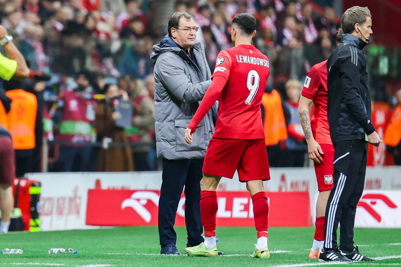 Jan Urban i Robert Lewandowski Trener w kurtce stoi na murawie i ściska dłoń piłkarza w czerwonym stroju z numerem 9, podpisanym 'Lewandowski'. W tle inni zawodnicy oraz widzowie na trybunach. Na boisku obecne są napisy reklamowe, atmosfera sportowa.