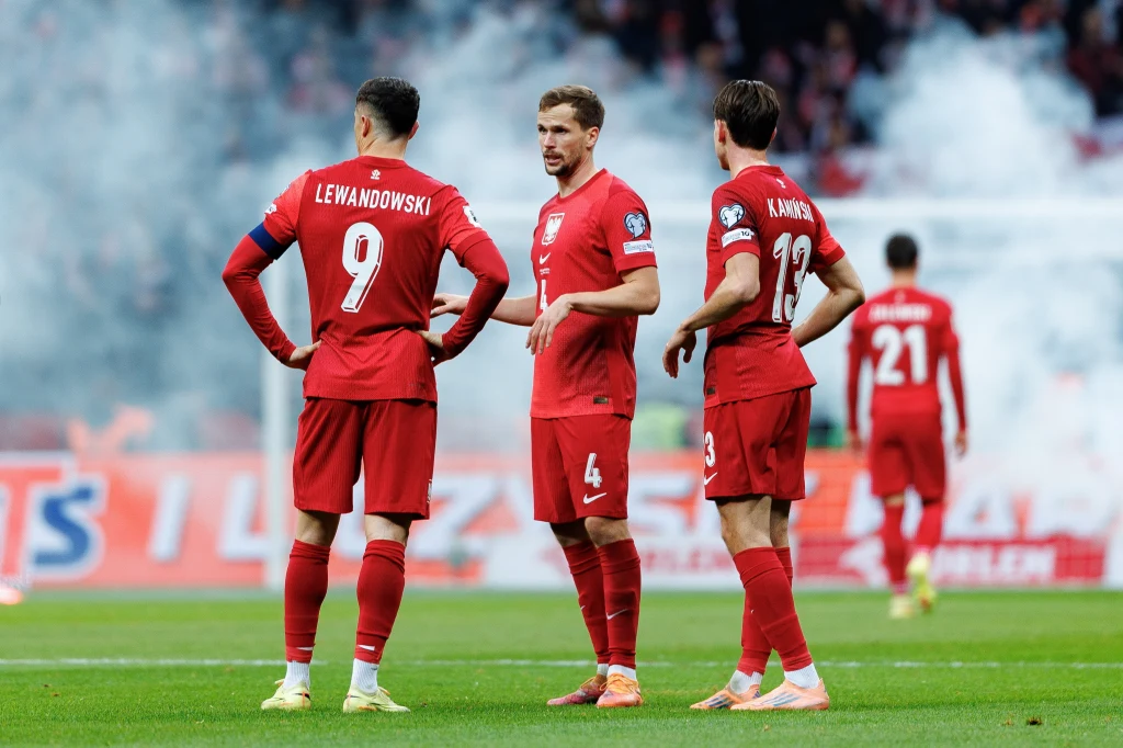 Piłkarze kadry Polski podczas meczu z Holandią Trzech polskich piłkarzy w czerwonych strojach narodowych stoi na murawie stadionu podczas meczu, w tle widoczni są inni zawodnicy oraz unoszący się dym z rac.