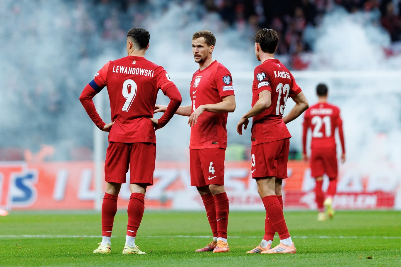 Piłkarze kadry Polski podczas meczu z Holandią Trzech polskich piłkarzy w czerwonych strojach narodowych stoi na murawie stadionu podczas meczu, w tle widoczni są inni zawodnicy oraz unoszący się dym z rac.
