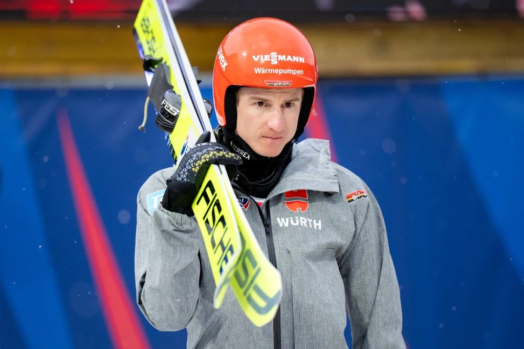 Zawodnik w kurtce sportowej i kasku narciarskim trzymający narty na ramieniu, na tle niebieskiej planszy i drewnianego elementu.