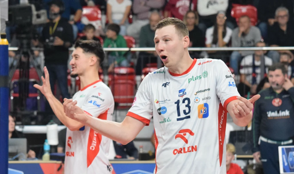 Rafał Szymura i jego koledzy sprawili, że ZAKSA Kędzierzyn-Koźle w końcu wygrała tie-breaka Dwóch siatkarzy w białych koszulkach z czerwonymi akcentami, z widocznymi numerami oraz logotypami sponsorów, wyraża emocje podczas meczu. W tle widać kibiców na trybunach.