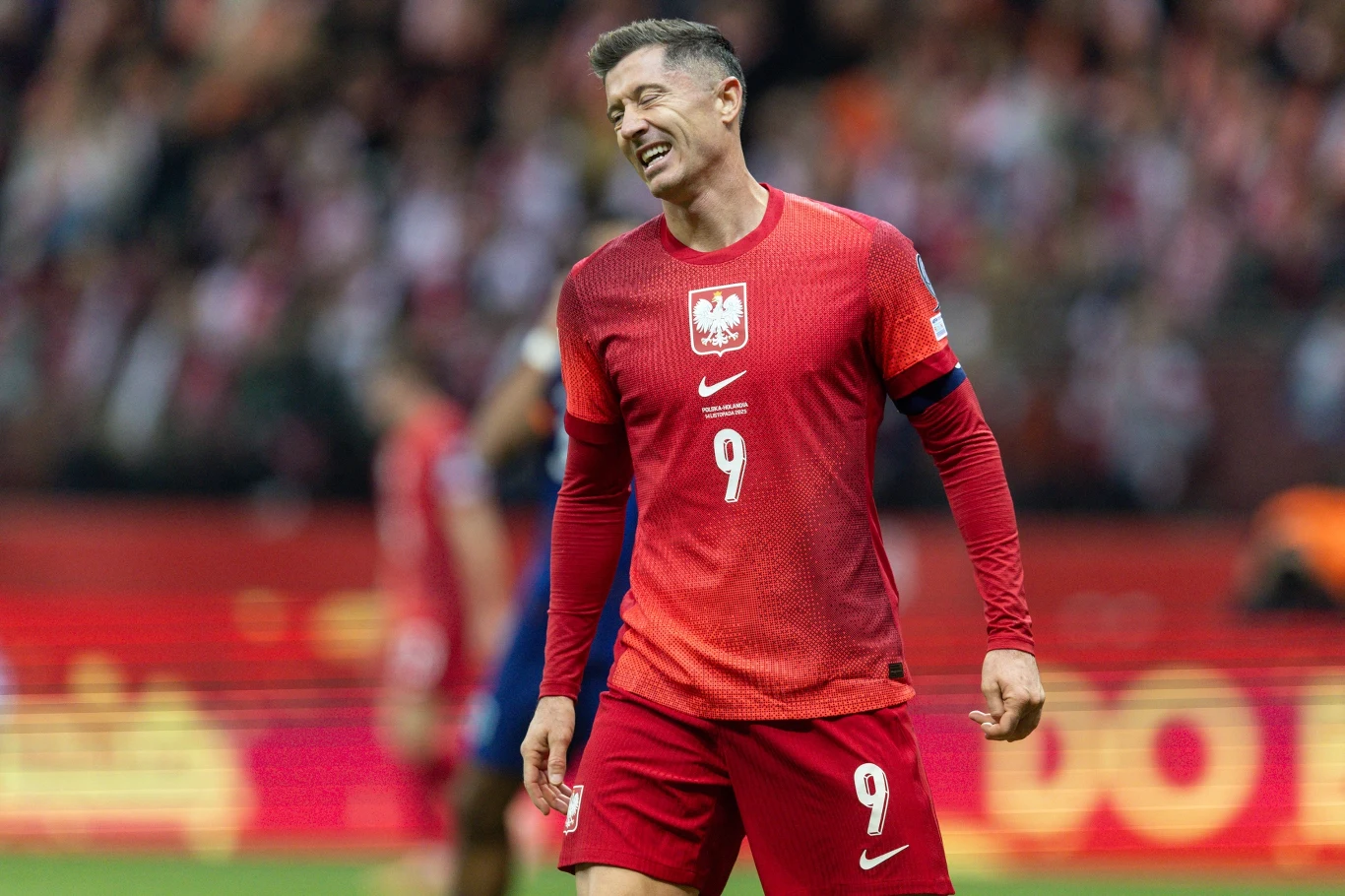 Piłkarz w czerwonym stroju reprezentacji Polski z numerem 9 na koszulce stoi na boisku z grymasem na twarzy. W tle rozmyta publiczność oraz fragmenty stadionu.