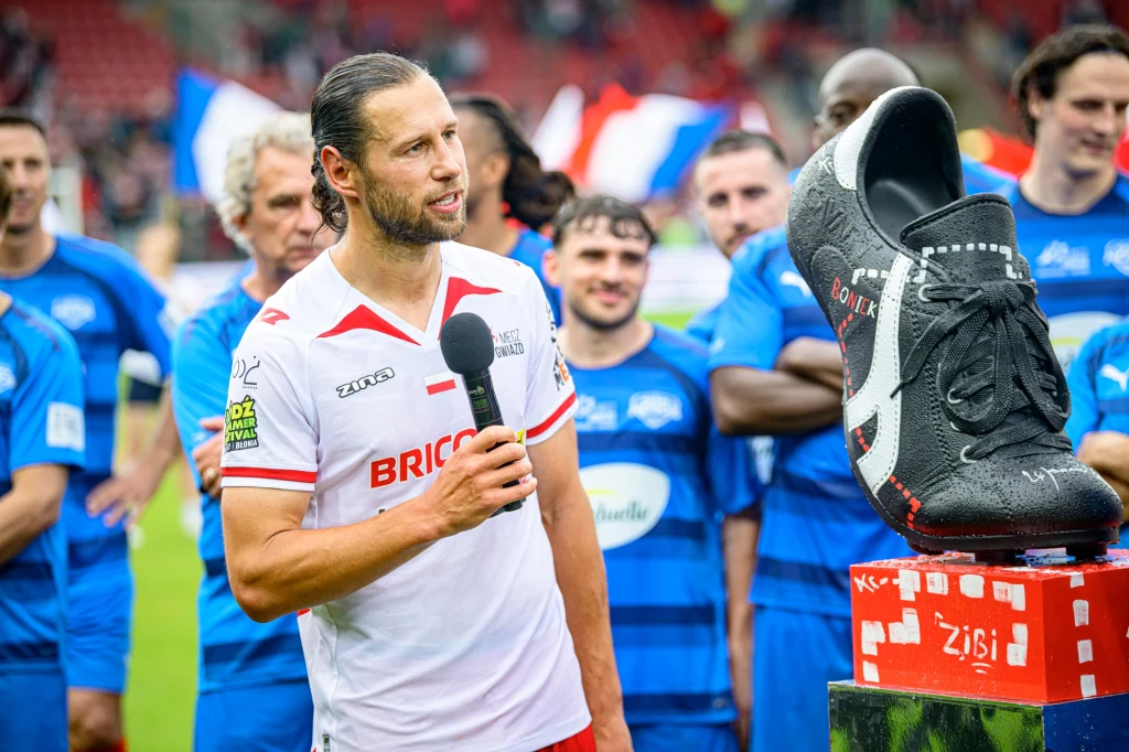 Piłkarz w białej koszulce z mikrofonem w ręku stoi przed wielkim modelem buta piłkarskiego umieszczonym na podeście. W tle grupa zawodników w niebieskich strojach i rozmazane flagi na trybunach stadionu.