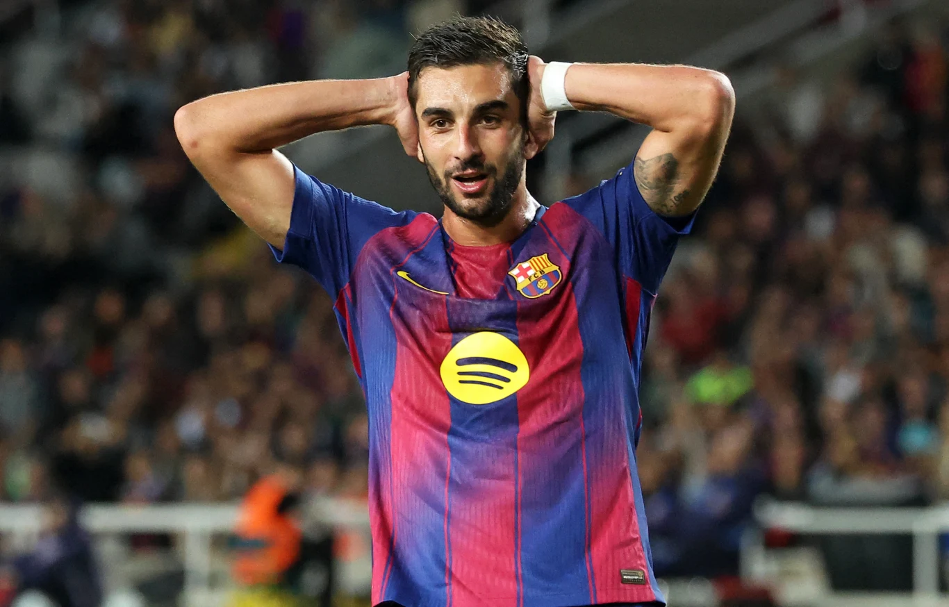 Piłkarz w koszulce FC Barcelony stoi na murawie z rękami założonymi za głową. W tle widać trybuny stadionu oraz sylwetki kibiców.