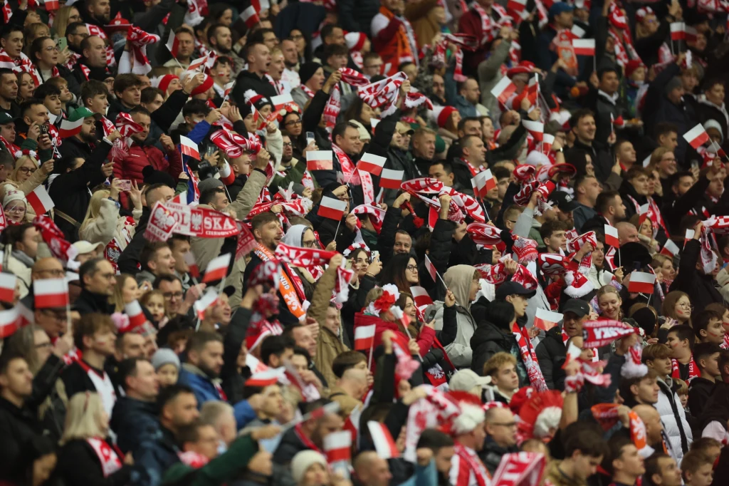 Tłum kibiców ubranych w biało-czerwone barwy oraz szaliki, unoszących ręce i świętujących wspólnie na trybunach stadionu sportowego.