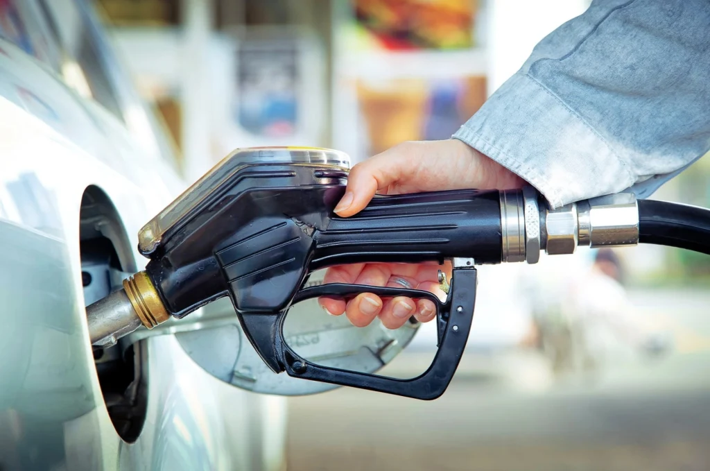 Ręka trzymająca pistolet dystrybutora paliwa w trakcie tankowania samochodu, fragment pojazdu widoczny na pierwszym planie.