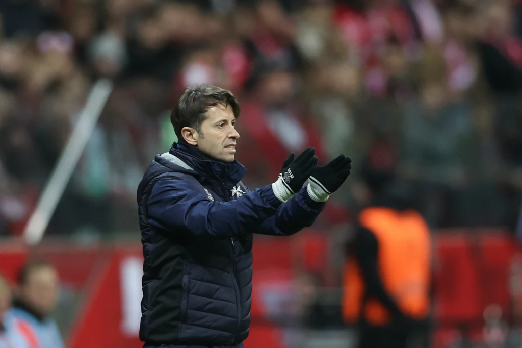 Trener w kurtce sportowej i rękawiczkach gestykuluje dłońmi, kibice w tle na trybunach stadionu skupieni na wydarzeniach boiskowych.