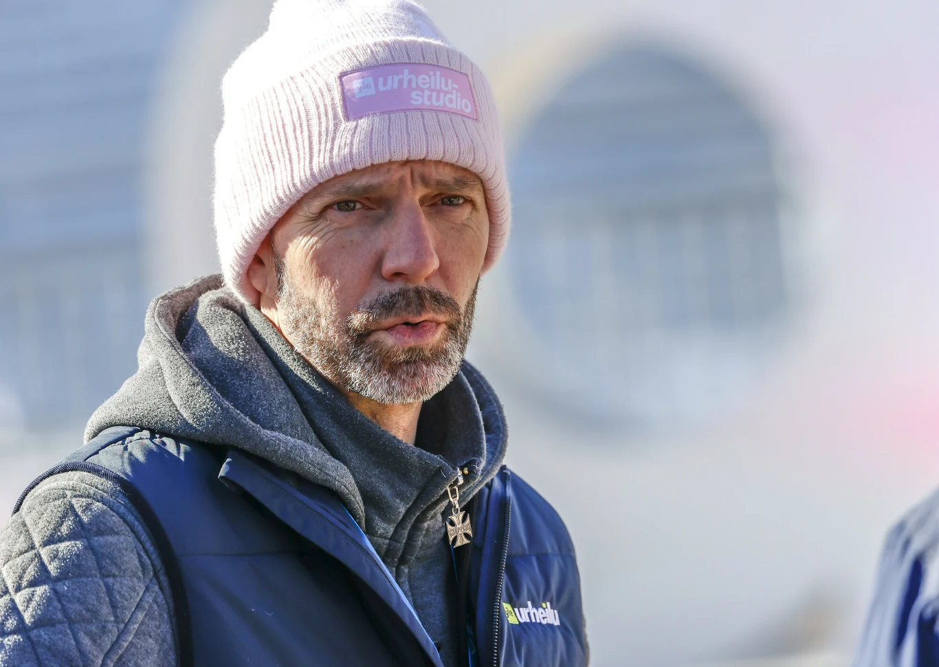 Janne Ahonen Mężczyzna w różowej czapce oraz niebieskiej kurtce na świeżym powietrzu, z lekkiem zarostem, tle rozmyte okrągłe kształty budynku.