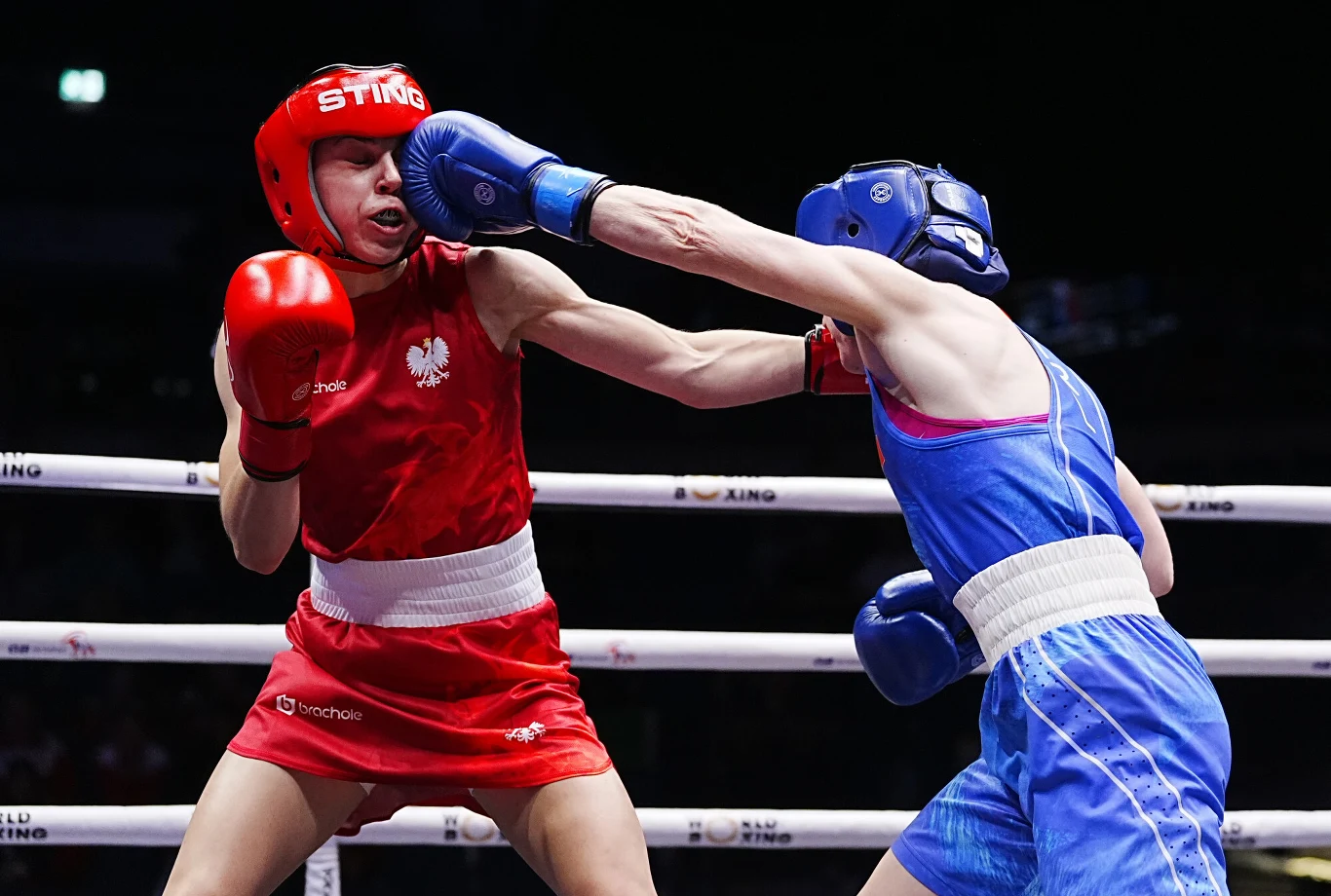 Dwóch bokserów w trakcie pojedynku na ringu, jeden w czerwonym stroju otrzymuje cios od rywala w niebieskim stroju, mocno skoncentrowani na walce.