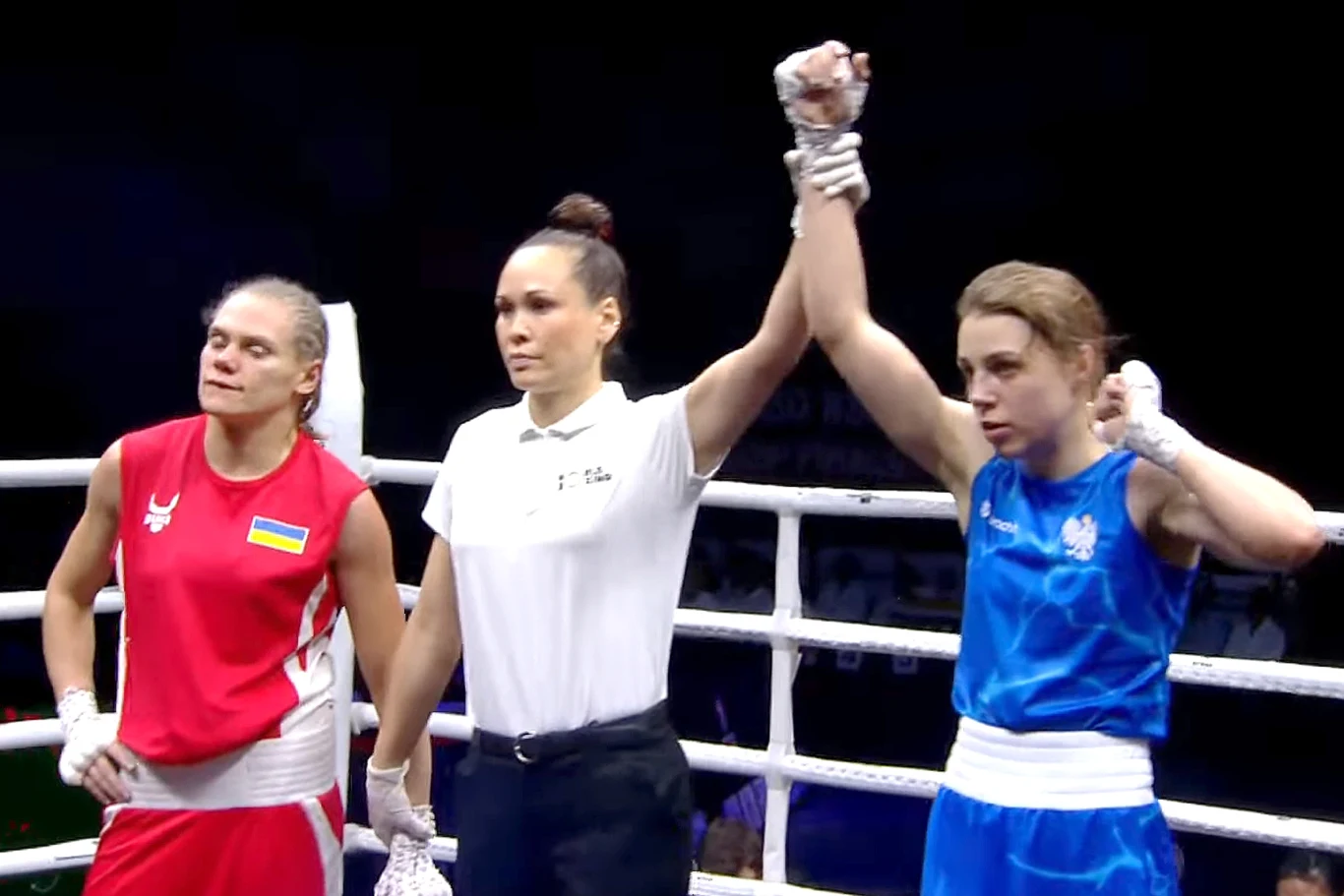 Sędzia w ringu podnosi rękę zawodniczki bokserskiej w niebieskim stroju jako gest zwycięstwa, podczas gdy zawodniczka w czerwonym stroju pozostaje obok z wyrazem przegranej.