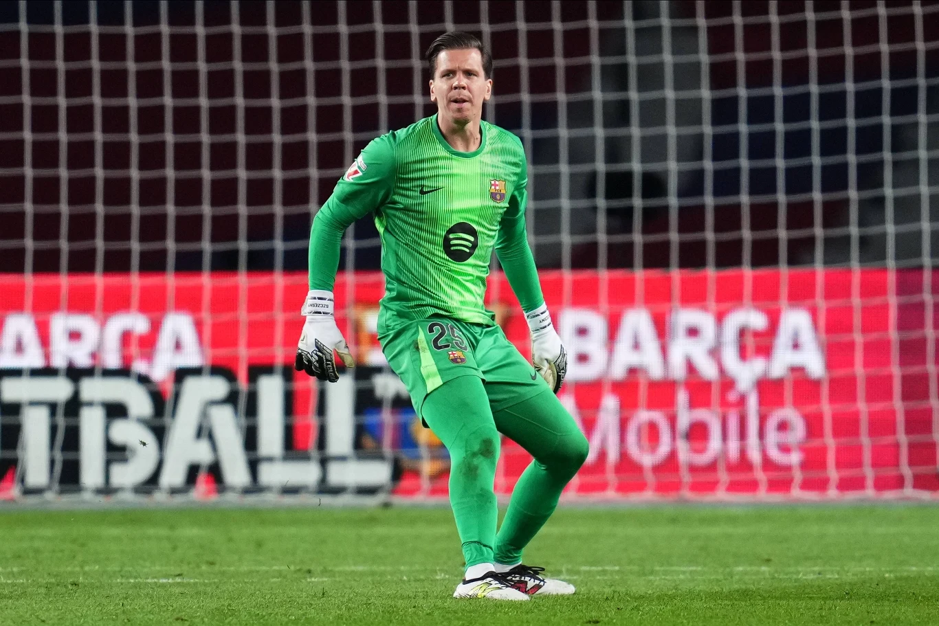 Bramkarz w zielonym stroju sportowym stoi na murawie stadionu, skoncentrowany i gotowy do interwencji w trakcie rozgrywanego meczu piłkarskiego. W tle widać bandy reklamowe z napisem Barca.