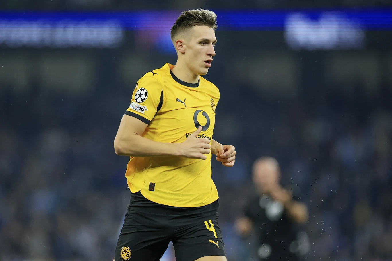 Piłkarz w żółtej koszulce Borussii Dortmund grający na boisku w trakcie meczu, z opaską Ligi Mistrzów na ramieniu, w tle rozmazany tłum i zawodnicy.