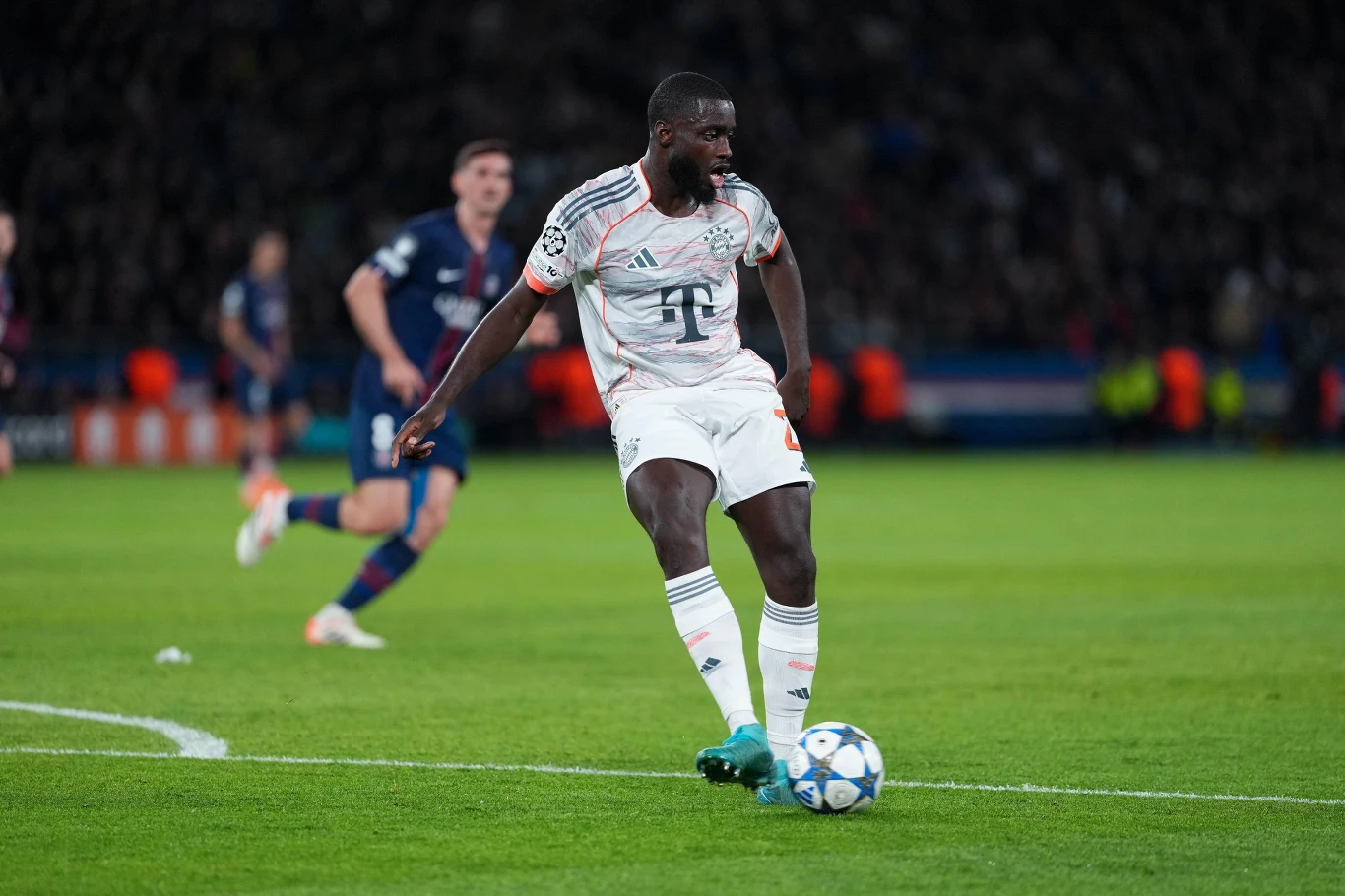 Piłkarz w stroju Bayernu Monachium kontroluje piłkę na boisku podczas meczu, w tle widoczni są zawodnicy drużyny przeciwnej oraz kibice na trybunach.