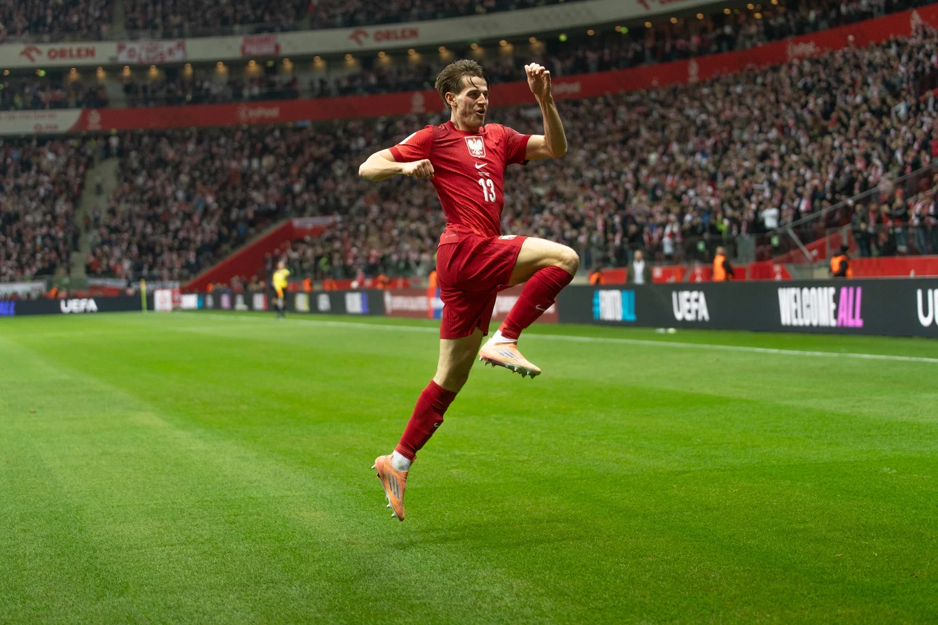 Jakub Kamiński Piłkarz w czerwonym stroju wykonuje energiczny skok z zaciśniętą pięścią w geście radości na tle wypełnionego kibicami stadionu.