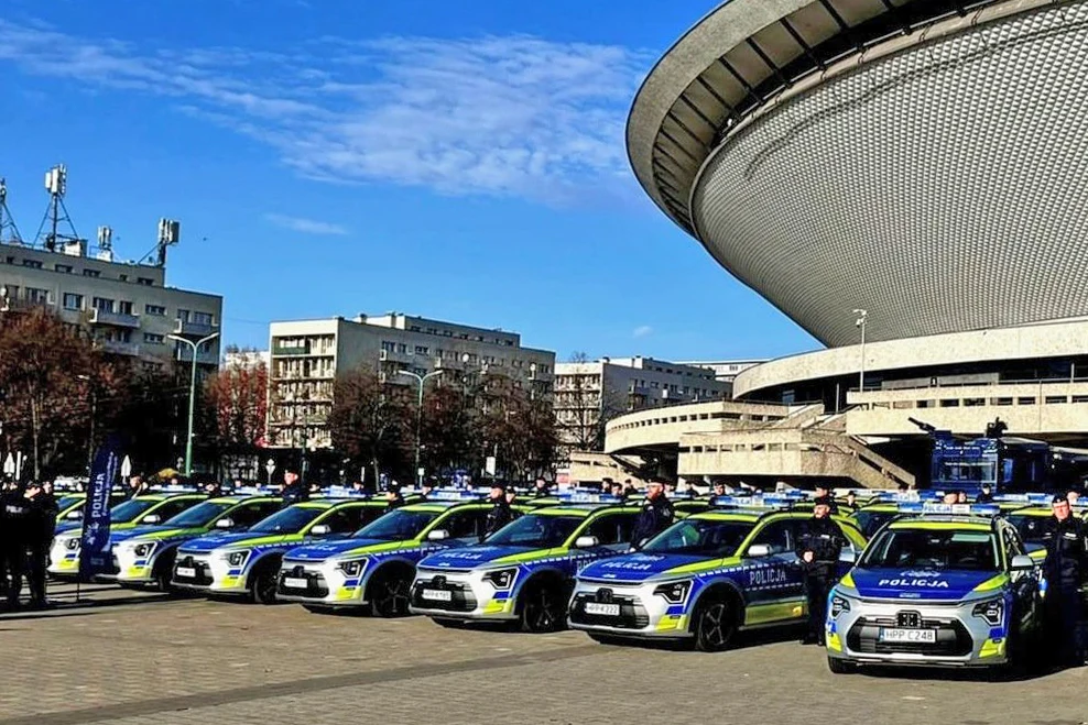 Szereg nowych radiowozów policyjnych ustawionych na placu przed charakterystyczną halą widowiskową Spodek w Katowicach, w tle bloki mieszkalne i niebieskie niebo.