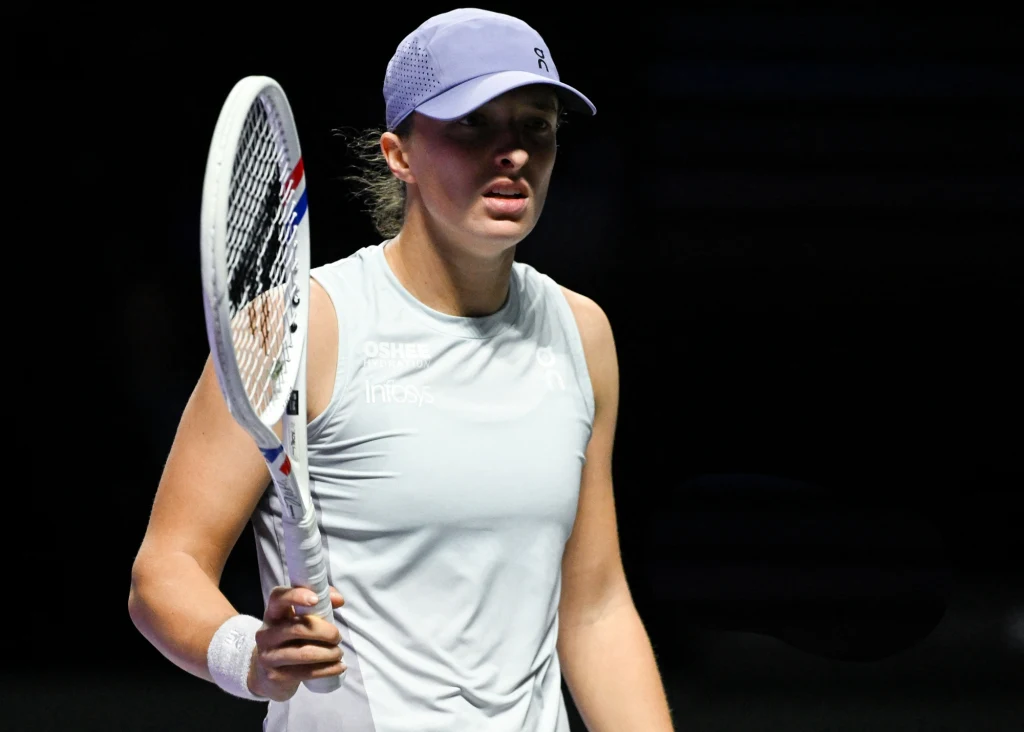 Iga Świątek Tenisistka w sportowej koszulce i czapce z daszkiem trzyma rakietę tenisową w ręku, na tle ciemnego boiska, wyrażając skupienie i determinację.