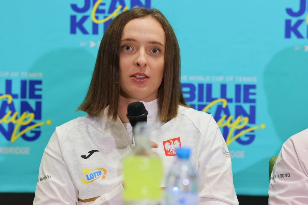 Iga Świątek podczas BJK Cup w Gorzowie Wielkopolskim Kobieta w białej sportowej kurtce z polskim godłem, siedząca przy mikrofonie na tle niebieskiego baneru z napisem 'Billie Jean King Cup', przed nią plastikowa butelka i żółto-zielony napój.