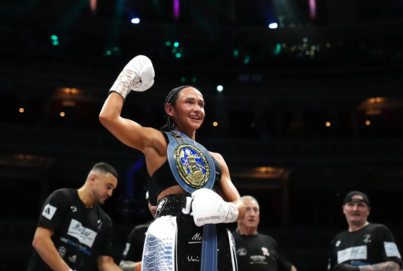 Zwyciężczyni walki bokserskiej w ringu z ręką uniesioną w geście triumfu, ubrana w sportowy strój i białe rękawice bokserskie, na jej ramieniu wisi ozdobny pas mistrzowski. W tle widoczni są członkowie jej zespołu.