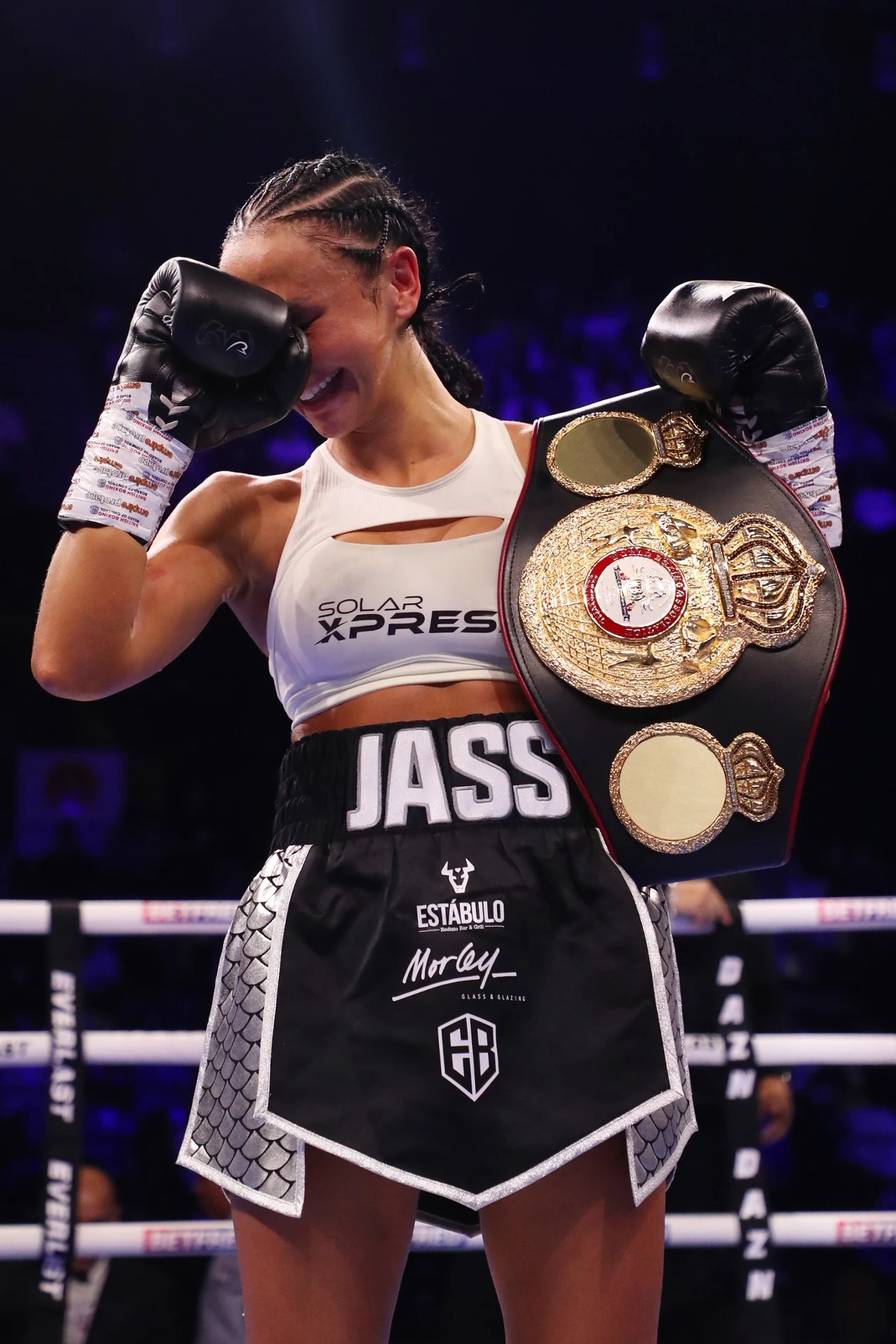 Kobieta ubrana w sportowy strój bokserski stoi w ringu z rękawicami bokserskimi, trzymając pas mistrzowski przewieszony przez ramię. Okazuje emocje, częściowo zasłaniając twarz dłonią. W tle widoczne rozmyte światła areny.