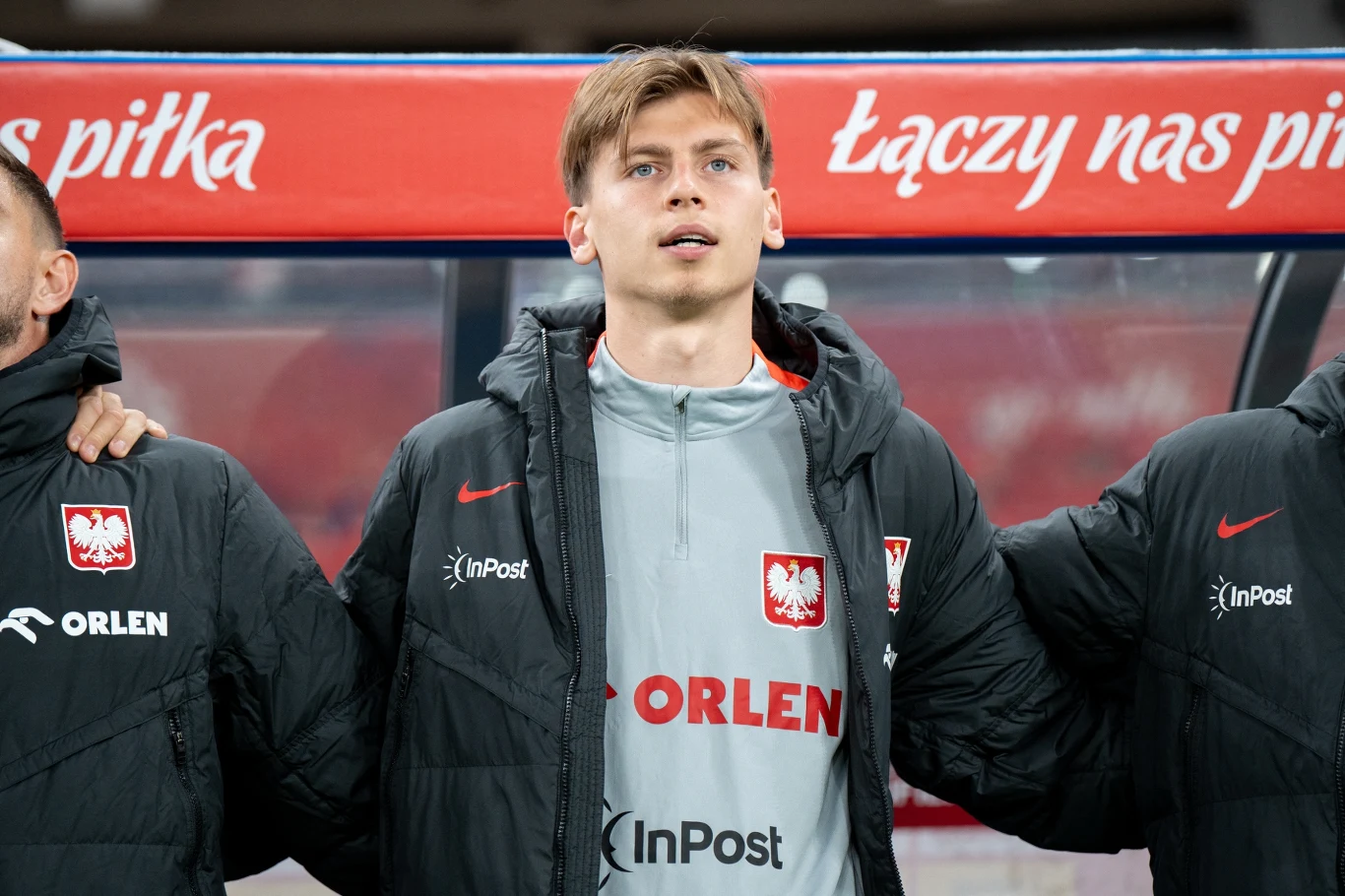 Młody mężczyzna w sportowej kurtce z emblematem reprezentacji Polski stoi na stadionie, otoczony przez innych zawodników, w tle widoczny jest fragment ławki rezerwowych oraz napis 'Łączy nas piłka'.