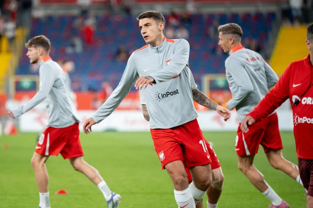 Jakub Kiwior Piłkarze w jasnoszarych koszulkach i czerwonych spodenkach wykonują ćwiczenia rozgrzewające na zielonym boisku stadionowym, w tle widoczna jest pusta część trybun.