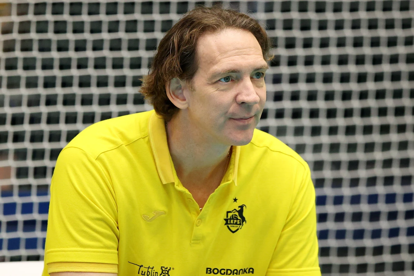 Mężczyzna w żółtej koszulce sportowej z logotypami sponsorów i klubu siatkarskiego, siedzący na tle siatki sportowej, skierowany lekko w bok, sprawiający wrażenie skupionego i spokojnego.