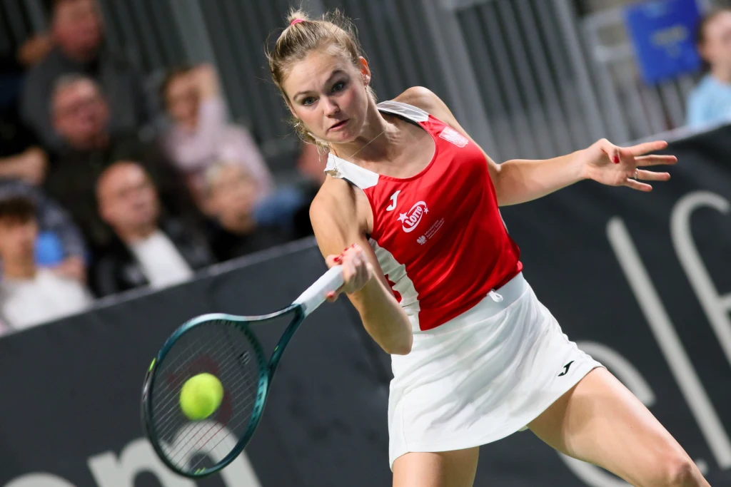 Tenisistka w czerwono-białym stroju sportowym wykonuje dynamiczne uderzenie rakietą podczas meczu tenisowego, w tle widoczna rozmazana publiczność na trybunach.