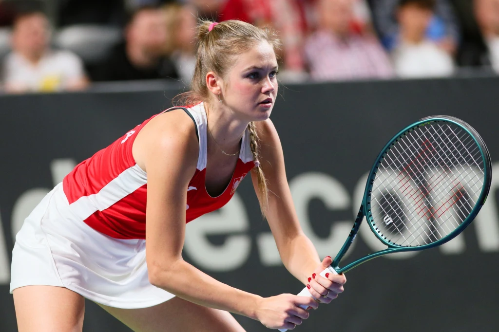 Tenisistka w czerwono-białym stroju przygotowuje się do odbioru piłki podczas meczu na korcie tenisowym, trzymając rakietę oburącz, skupiona na grze