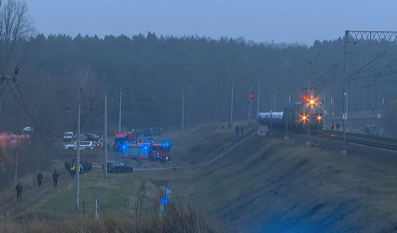 Służby ratunkowe i policyjne pracujące w pobliżu torów kolejowych, kilka pojazdów uprzywilejowanych z włączonymi światłami, zbliżająca się lokomotywa, ludzie rozproszeni wokół miejsca zdarzenia, las w tle.