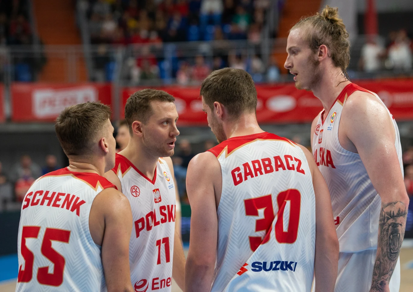 Czterech koszykarzy reprezentacji Polski w białych strojach z czerwonymi napisami stoi blisko siebie na parkiecie podczas meczu, prowadząc intensywną rozmowę, w tle widoczna zamazana widownia i elementy hali sportowej.