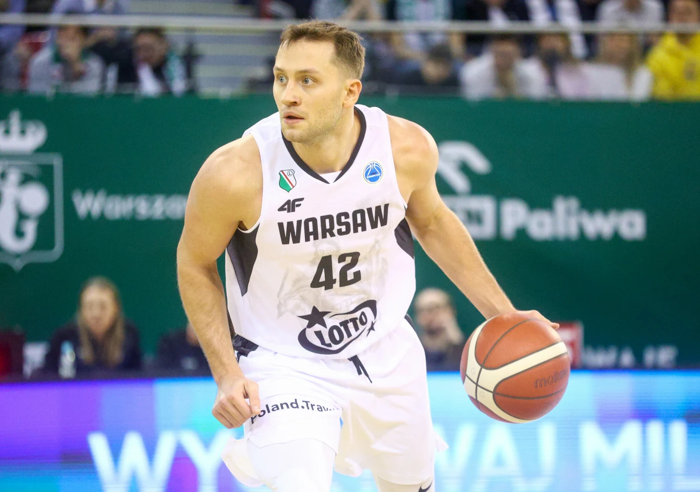 Koszykarz w białym stroju drużyny Warszawa z numerem 42 prowadzi piłkę w trakcie meczu na hali sportowej. W tle widoczni kibice oraz banery reklamowe.
