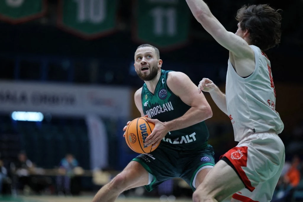 Koszykarz w zielonym stroju z napisem Wrocław przygotowuje się do rzutu piłką do kosza, intensywnie obserwując sytuację na boisku. Obrońca w białym stroju próbuje go zablokować, podnosząc rękę. W tle rozmazana publiczność i fragment hali.
