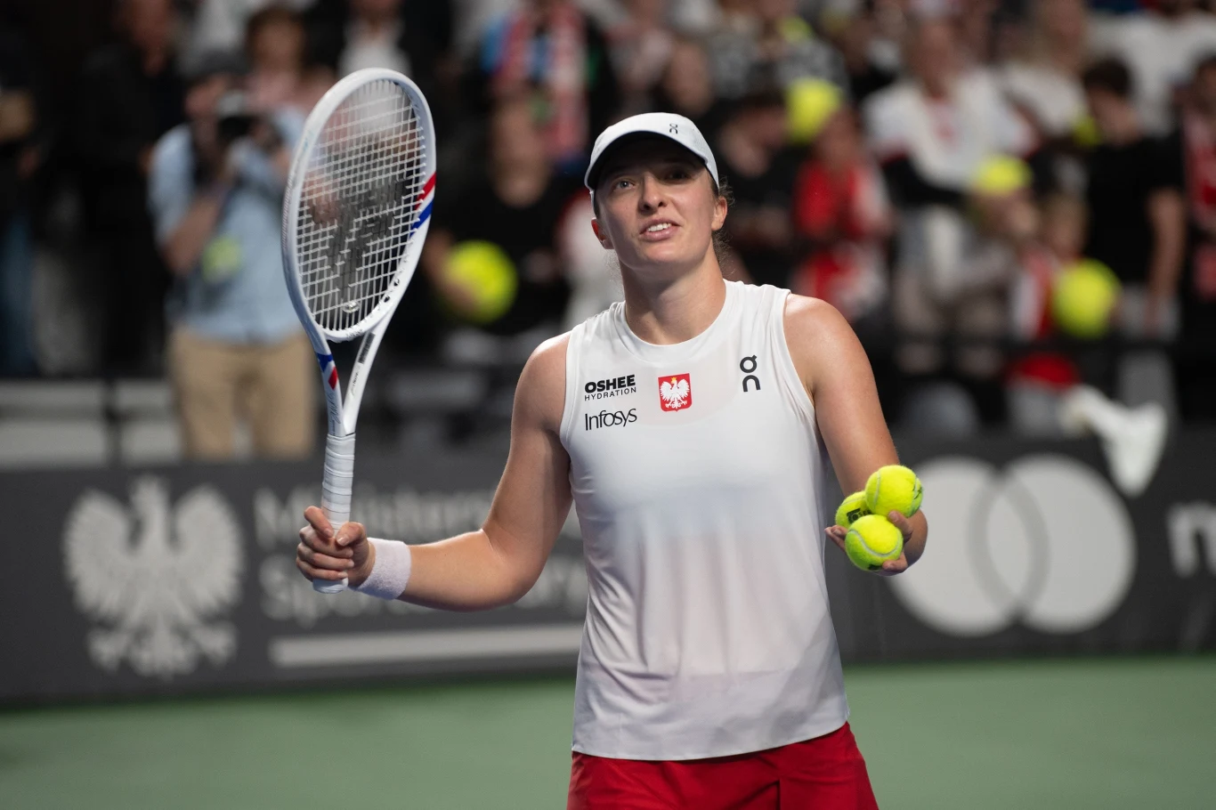 Tenisistka w białej czapce i stroju sportowym trzymająca rakietę tenisową i dwie piłki na tle kortu, w otoczeniu kibiców i polskich symboli narodowych.